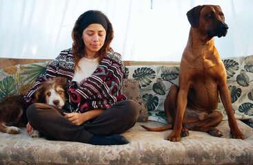 A young woman sits cross-legged on a couch, wearing a patterned sweater, with one dog resting beside her and another larger dog sitting attentively nearby.
