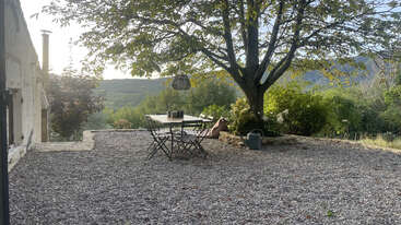 A peaceful outdoor patio with a gravel ground, metal table and chairs under a large shady tree. Scenic views of distant hills and lush greenery surround.
