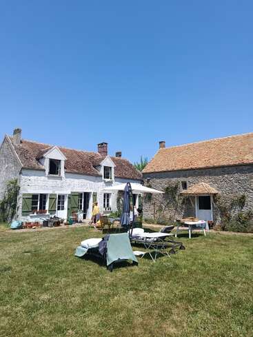 Ein charmantes rustikales Haus mit grünen Fensterläden und einem steinernen Nebengebäude steht unter einem klaren blauen Himmel, umgeben von grünem Rasen und Gartenmöbeln. Friedliche Atmosphäre.