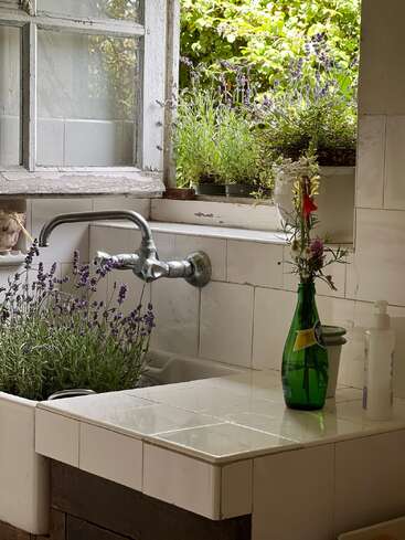 Sonnenlicht strömt durch ein offenes Fenster über einem rustikalen Spülbecken, das mit Lavendelpflanzen geschmückt ist. Eine grüne Flasche mit Wildblumen verleiht dem Ganzen einen natürlichen Charme. Eine friedliche, gemütliche Atmosphäre.