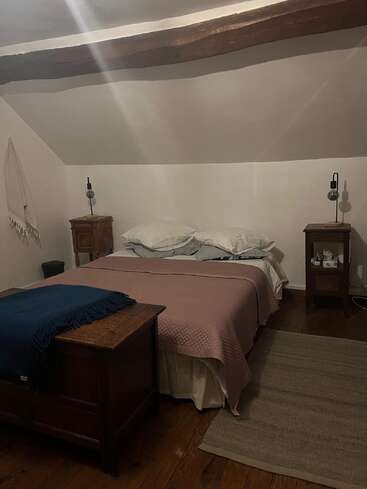 Dieses gemütliche Schlafzimmer verfügt über ein großes Bett mit rosa Bettwäsche, Holzmöbel, Nachttische mit Lampen, freiliegende Deckenbalken, schräge weiße Wände und einen Teppich.