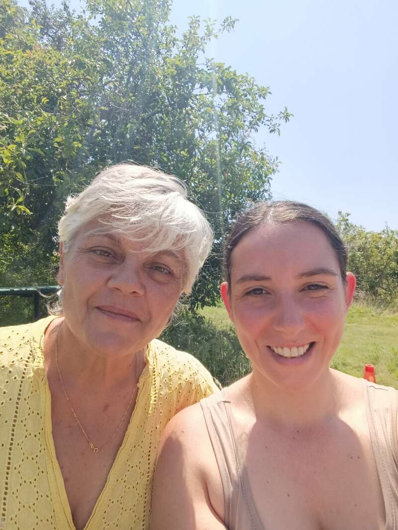Zwei Frauen lächeln an einem sonnigen Tag im Freien. Die eine hat kurzes graues Haar und trägt ein gelbes Oberteil; die andere hat dunkles Haar und ein hellbraunes Oberteil. Die Bäume dahinter.