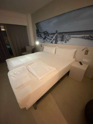 L'image représente une chambre d'hôtel avec deux lits, chacun avec des draps et des oreillers blancs, sur fond d'une grande photographie en noir et blanc accrochée au mur.