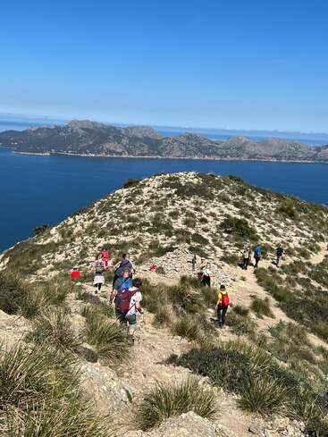 Un groupe de randonneurs gravit une colline rocheuse et herbeuse surplombant une mer d'un bleu magnifique, avec des montagnes escarpées et un ciel clair en toile de fond.