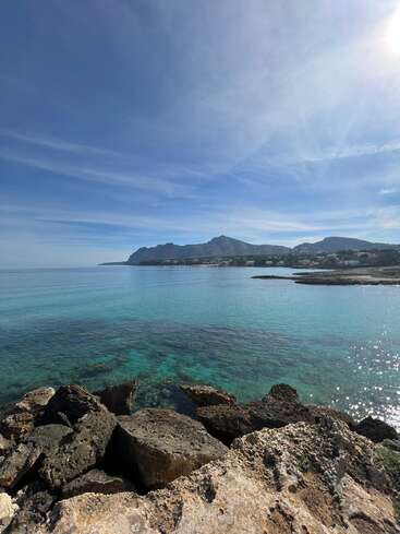 Une eau turquoise cristalline scintille près des côtes rocheuses, tandis que les montagnes se dressent en arrière-plan sous un ciel bleu éclatant. Les reflets du soleil créent une scène côtière paisible.