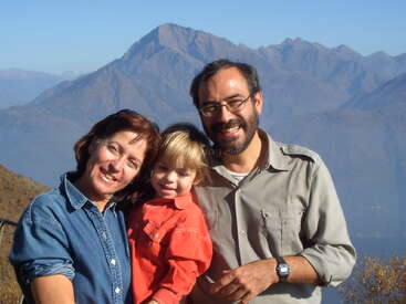 Une famille souriante de trois personnes pose en plein air. Des montagnes majestueuses et un ciel bleu clair forment un arrière-plan étonnant, capturant un moment de joie dans un environnement naturel magnifique.