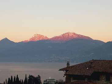 Les montagnes enneigées s'illuminent de rose au lever du soleil, surplombant un lac paisible et les toits d'un village avec des cheminées, entourés de collines et d'arbres sombres et ombragés. Atmosphère sereine.