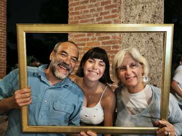Un homme souriant, une jeune femme et une femme plus âgée posent étroitement ensemble, tenant un cadre photo vide devant eux, sur fond de mur de briques et de pierres.