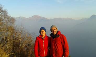 Un couple souriant, vêtu d'une veste rouge et d'un chapeau en tricot, pose en plein air. Des arbres dénudés et des montagnes lointaines enveloppées de brume constituent un arrière-plan naturel magnifique et serein au coucher du soleil.