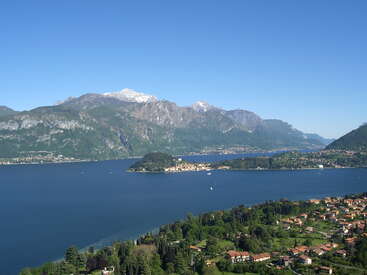 Uma bela cena de lago com águas azuis profundas, uma pequena vila em uma ilha, vegetação exuberante, casas distantes e montanhas imponentes com picos nevados sob um céu claro.