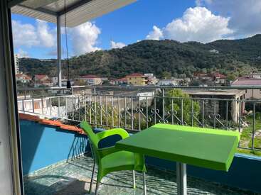 A green chair and table sit on a balcony overlooking a scenic town, green hills, scattered houses, blue sky, lush trees, and fluffy white clouds.