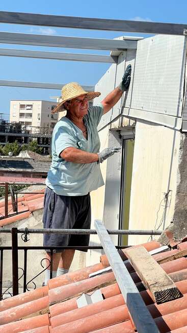 A man wearing a straw hat, gloves, and casual clothes works on a rooftop under a sunny sky, smiling while handling wall panels or construction materials.