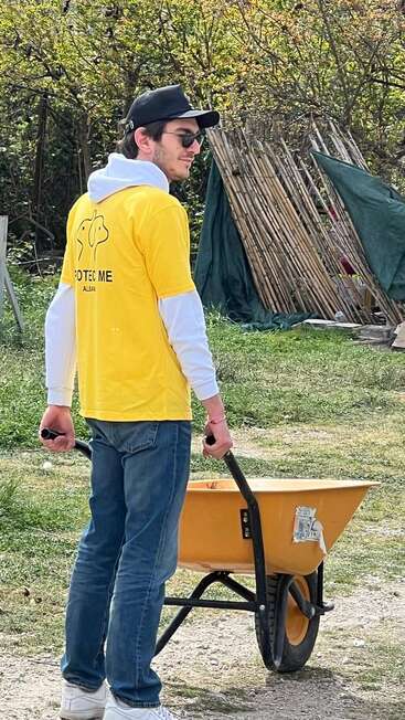 A young man wearing sunglasses, a yellow shirt, cap, and jeans is pushing a yellow wheelbarrow outdoors, surrounded by greenery and construction materials.