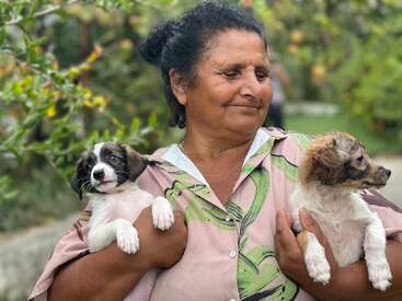 A woman stands outdoors, smiling warmly while holding two adorable puppies in her arms. Lush greenery surrounds them, creating a peaceful and heartwarming scene.