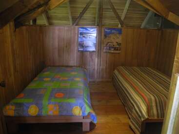 Esta acogedora habitación de madera cuenta con dos camas individuales con mantas de colores, techo inclinado, carteles en la pared del fondo y un ambiente cálido y rústico ideal para relajarse.