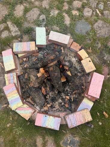 A small fire burns in a circular fire pit made of bricks colored with pastel chalk. The fire pit sits on grass and stone ground.