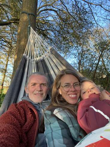Uma família feliz de três pessoas, sentada bem juntinha numa rede ao ar livre, sorri calorosamente para a câmera. As árvores e a luz do sol criam um cenário sereno e natural.
