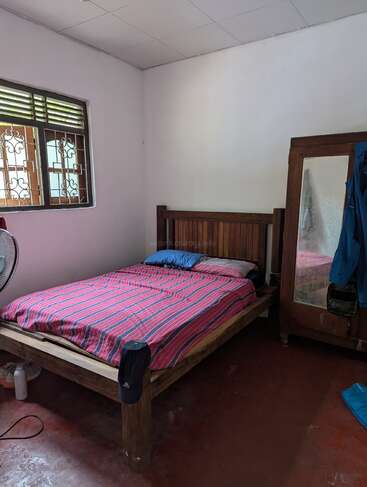 Um quarto simples com uma cama de madeira, roupa de cama listrada de vermelho, guarda-roupa com espelho, casaco azul pendurado, janela com grade, ventilador elétrico e alguns itens no chão.