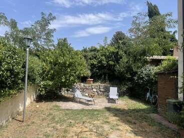 A peaceful backyard with two lounge chairs, surrounded by lush greenery and trees, a bicycle, a small wooden shed, stone wall, and a streetlamp under a blue sky.