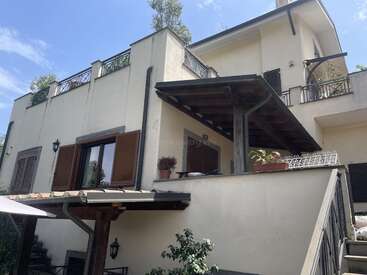 This image shows a light-colored, multi-story house with wooden shutters, balconies, and a slanted roof. There are potted plants, stair railings, and outdoor lights.
