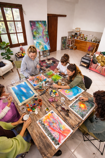 Cinq personnes sont assises autour d'une table dans une pièce chaleureuse et décorée d'œuvres d'art ; elles s'adonnent à la peinture d'œuvres abstraites aux couleurs vives à l'aide d'aquarelles et de divers matériaux artistiques. L'ambiance est créative.