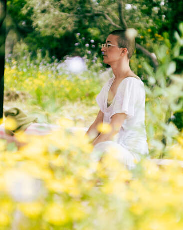 Une personne aux cheveux courts et portant des lunettes, vêtue de blanc, médite paisiblement en plein air, au milieu de fleurs d'un jaune éclatant et d'un feuillage verdoyant, créant ainsi une scène sereine et tranquille.