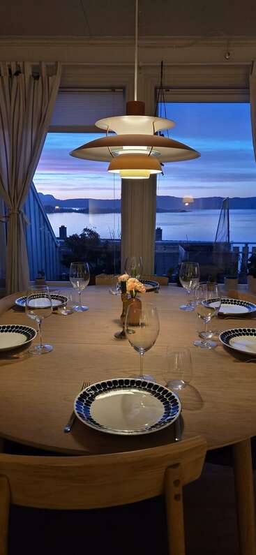 A beautifully set round dining table with six plates and wine glasses, elegant lamp above, large windows showing a stunning lake and mountain sunset view.