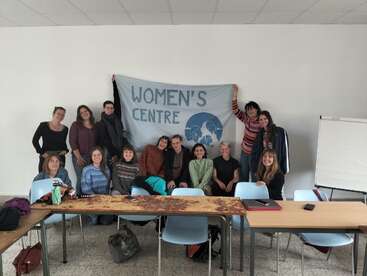Un grupo de trece personas posa en una sala, sonriente, con una pancarta del "Centro de la Mujer" detrás. Están sentadas y de pie alrededor de mesas, lo que sugiere camaradería.