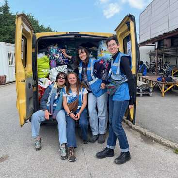 Four smiling women in blue vests pose in front of a yellow van filled with bags and supplies, suggesting teamwork, volunteering, and a positive, cooperative spirit.