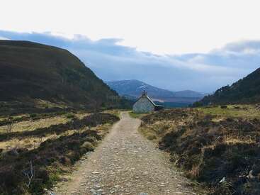 A rocky path leads through a rugged valley towards a small stone cottage. Hills rise on both sides, with distant snow-capped mountains under a cloudy sky.