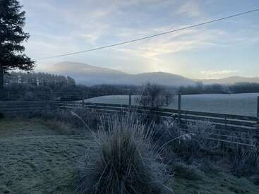 A frosty winter morning landscape with misty hills, pale blue sky, fences, frosted grass, trees, and distant mountains under soft early sunlight. Peaceful, serene scene.