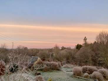 A frosty countryside landscape at sunrise, with a small stone house, bare trees, and colorful sky gradients. The scene feels tranquil and cold, inviting peace.