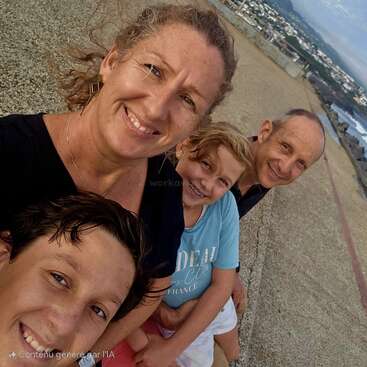 Uma família sorridente de quatro pessoas tira uma selfie em close-up ao ar livre. Atrás deles, há uma vista panorâmica com água, prédios urbanos distantes e montanhas sob um céu nublado.