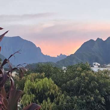 Um belo pôr do sol ilumina o céu com suaves tons de rosa e laranja, atrás de montanhas impressionantes, árvores verdes exuberantes e telhados espalhados em uma paisagem serena.