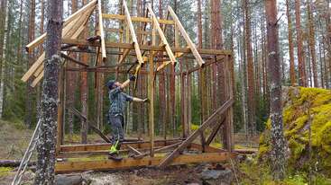 A person is constructing a wooden structure in a forest, attaching roof beams to the frame, surrounded by tall pine trees, rocks, and green moss.
