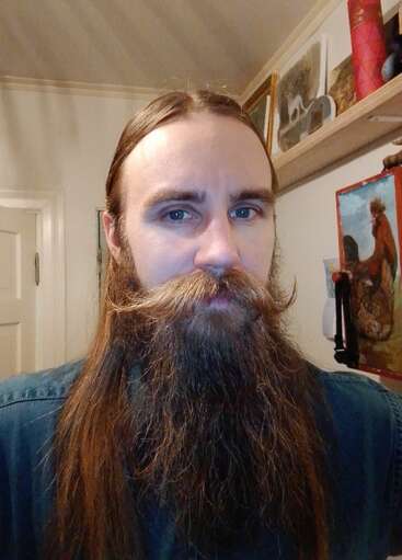 Un hombre con el pelo largo y castaño y una barba espesa y tupida está en el interior. Su bigote está peinado hacia arriba. Hay cuadros y estanterías al fondo.