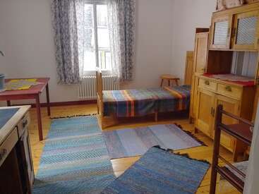 La imagen muestra una habitación luminosa y sencilla con una cama individual, alfombras tejidas de colores, muebles de madera, una mesa, una ventana con cortinas y luz natural.