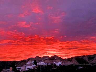 A breathtaking sunset illuminates the sky with vivid shades of red, pink, and purple above a small, peaceful village nestled among rolling hills and mountains.