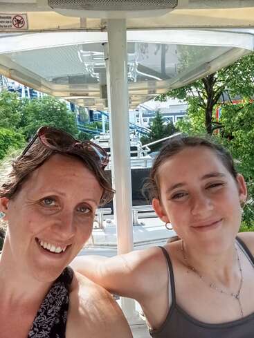 Two women smile for a selfie on an amusement park ride. Sunlight shines, trees and roller coaster tracks visible in the background. Happy, relaxed summer day.