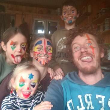 Un groupe joyeux de cinq personnes, le visage peint de couleurs vives, souriant et faisant des grimaces, semble s'adonner ensemble à une activité ludique et créative à l'intérieur.