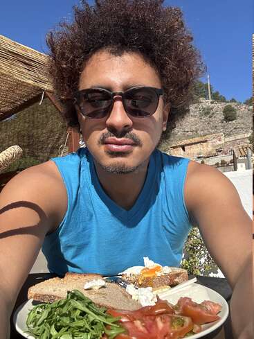 Um cara de óculos escuros e camiseta azul sem mangas tira uma selfie ao ar livre, aproveitando um dia ensolarado com um prato de torradas, ovos, rúcula e tomates.