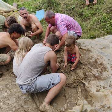 Das Bild zeigt eine Gruppe von Menschen, darunter auch Kinder, die gemeinsam an einem schlammigen Projekt auf einer Wiese arbeiten, während ein großes Stück Plastikfolie auf dem Boden liegt.