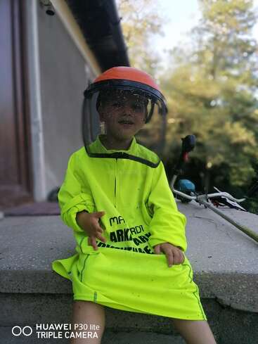 Un jeune enfant est assis sur des marches en béton ; il porte un gilet de sécurité jaune vif trop grand pour lui et un masque de protection orange, et sourit à l'extérieur avec de la verdure en arrière-plan.