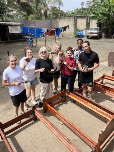 A group of seven smiling people are outdoors, painting wooden bed frames. Laundry hangs in the background, and tropical plants surround the dusty, community space.