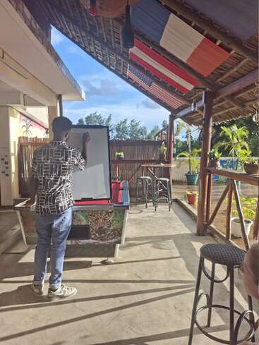 A man writes on a whiteboard in a bright outdoor area with barstools, flags above, plants around, and sunlight streaming through the wooden roof structure.