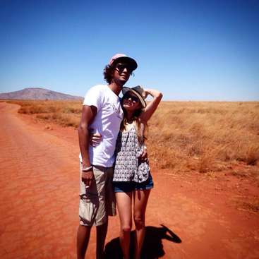 A imagem mostra um homem e uma mulher em pé em uma estrada de terra em uma paisagem desértica, com a mulher usando um chapéu e o homem com o braço em volta dela.