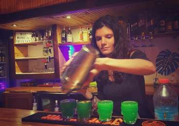 Uma mulher está despejando uma bebida de uma coqueteleira em uma fileira de bebidas verdes em um balcão de bar, com garrafas atrás dela.