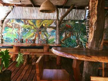A imagem mostra uma área de estar externa com tema tropical, com mesas, bancos e cadeiras de madeira, com um mural de animais e plantas da selva na parede.