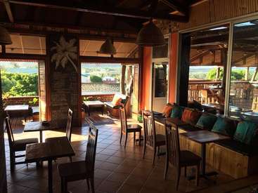 A imagem mostra uma sala espaçosa com móveis de madeira, com mesas e cadeiras, um sofá e uma grande janela com vista para o exterior e para as montanhas distantes.