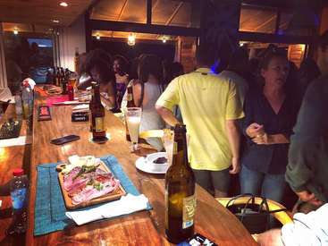 A imagem retrata uma cena de bar movimentada, com um balcão de madeira com bebidas e alimentos, cercado por clientes que se divertem em uma atmosfera animada.